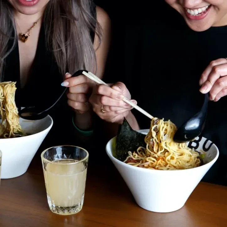 Two people eating ramen from white bowls using chopsticks and spoons.