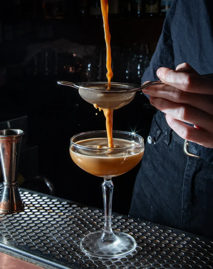 Bartender pouring golden cocktail through a strainer into a coupe glass.