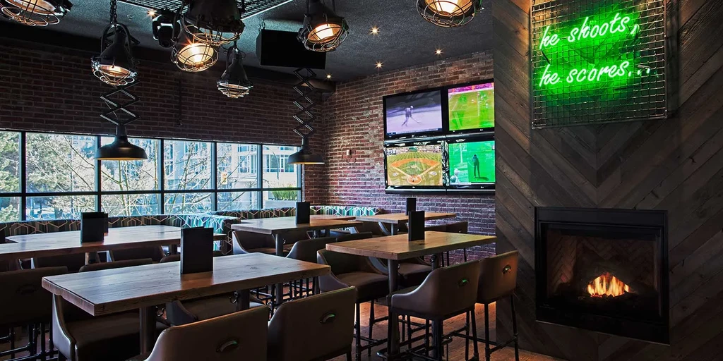 Empty industrial-chic restaurant with sports screens, fireplace, and neon sign.