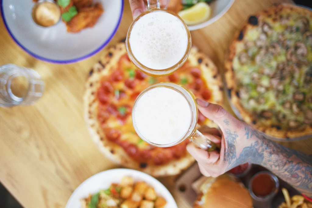 Two tattooed hands clinking beer mugs over a table of diverse food.