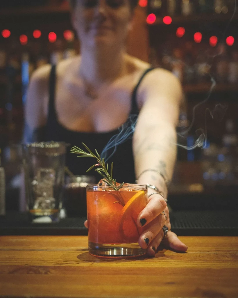 Hand holding smoky amber cocktail with orange and rosemary.