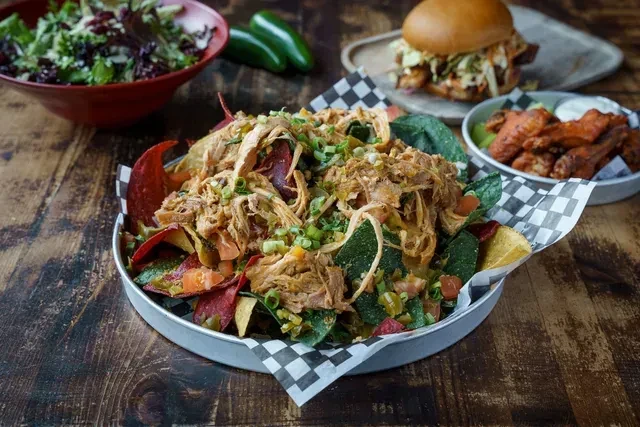 Colorful loaded nachos, salad, burger, and chicken wings on a table.