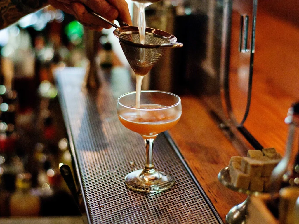 Bartender's hand straining an amber cocktail into a coupe glass.