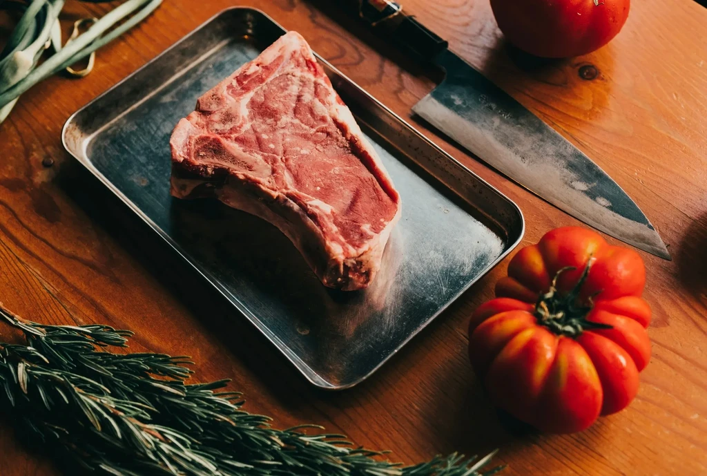 Raw T-bone steak, knife, tomatoes, rosemary, and spring onions on wood.