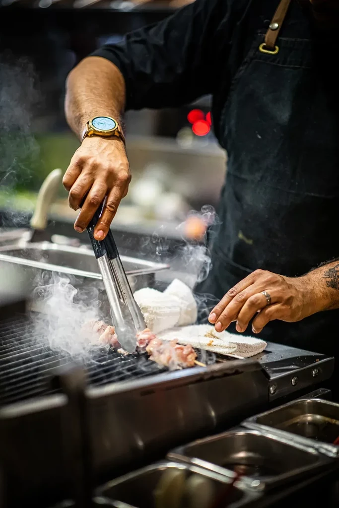 Chef's hands grilling meat skewers with tongs on a smoky grill.
