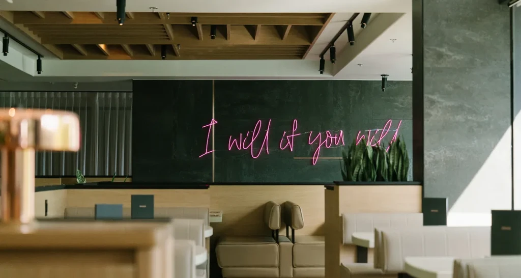 Modern restaurant interior with pink neon sign, booths, and plants.