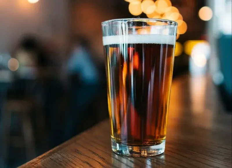 Glass of amber beer with foam on wood, blurred bokeh background.