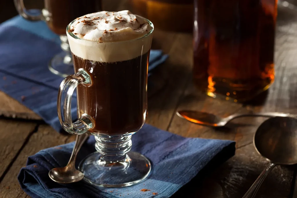 Glass mug with dark beverage, whipped cream, spoon on wood.