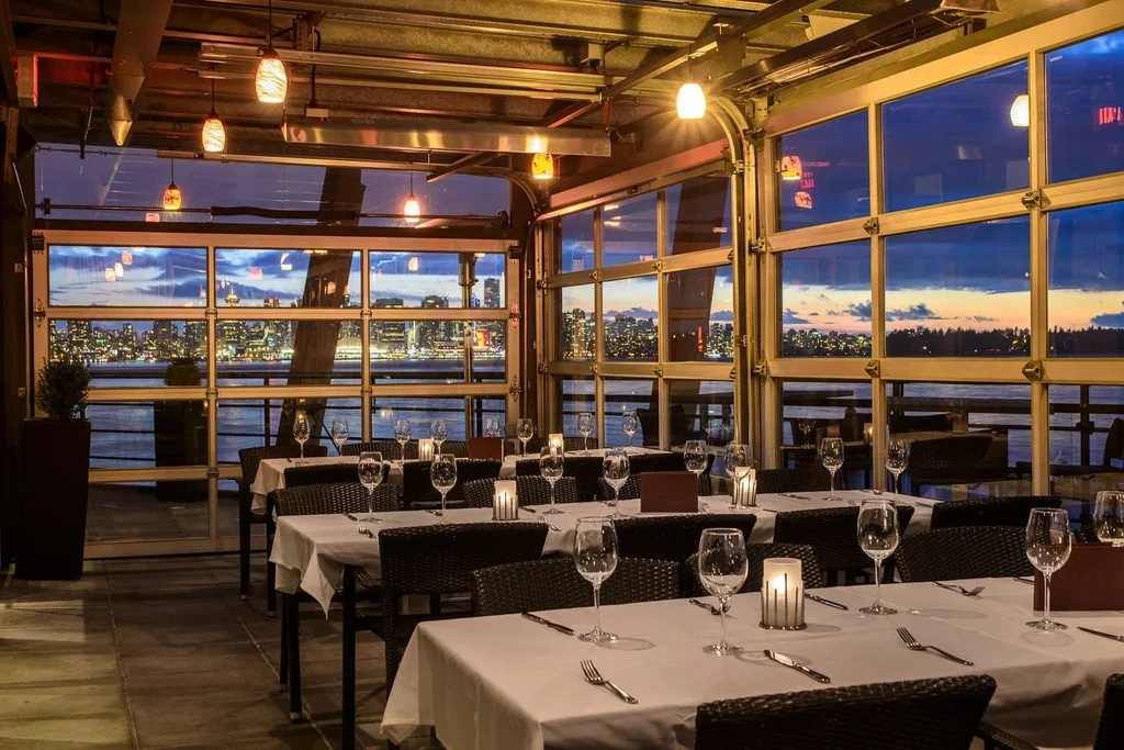 Restaurant interior, set tables, large windows, panoramic city view at dusk.