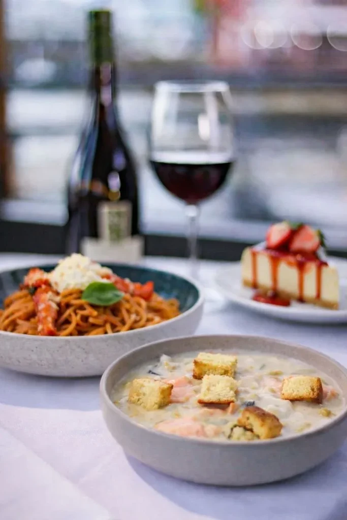 Table with creamy soup, seafood spaghetti, strawberry cheesecake, and red wine.