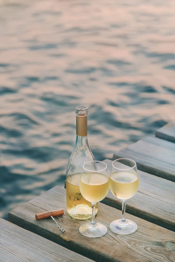 White wine bottle, two glasses, corkscrew on a wooden dock by water.