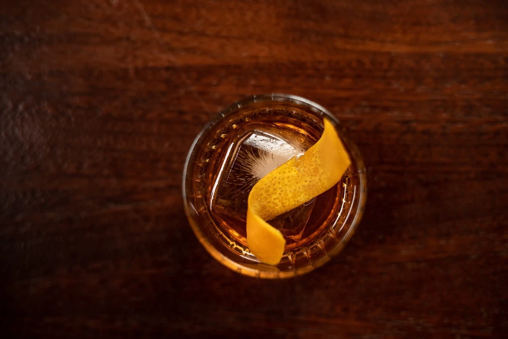Amber cocktail with orange peel and large ice cube on dark wood.