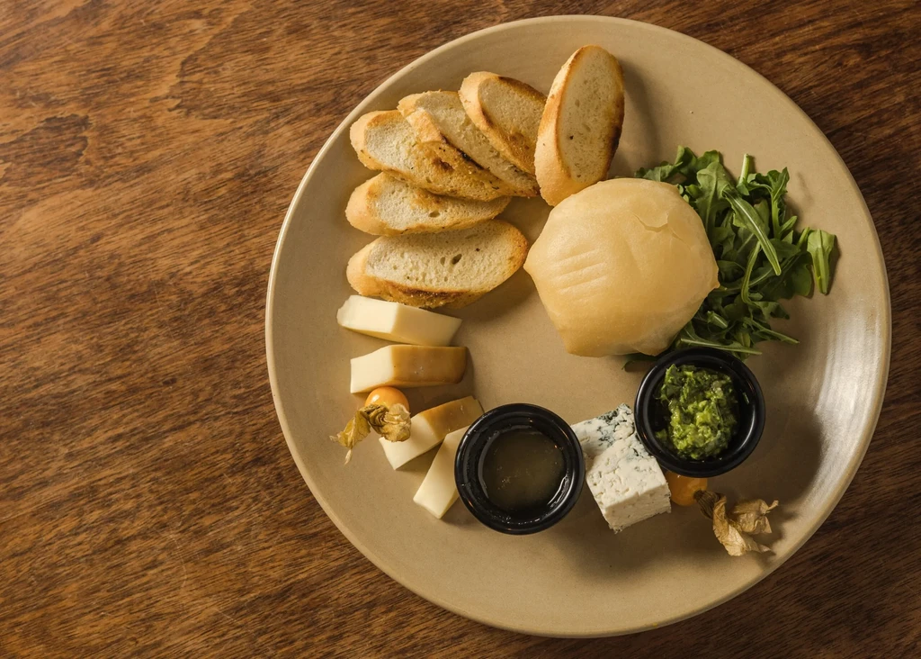 Top-down cheese board with bread, cheeses, and dips on wood.