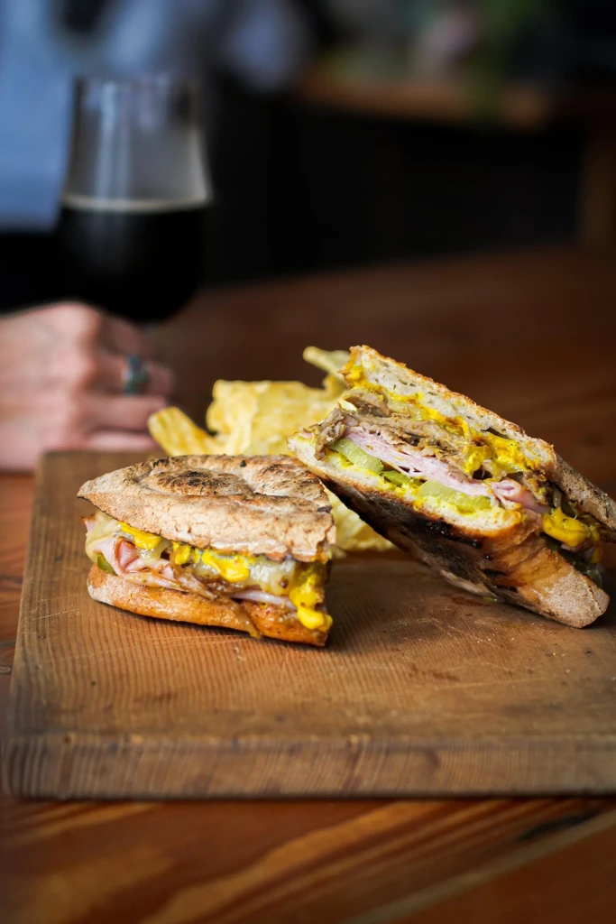 Pressed Cuban sandwich, cut in half, with chips and a beverage.