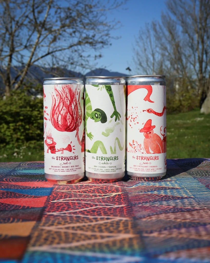 Three distinct 'the STRANGERS' wine cans on patterned fabric outdoors.