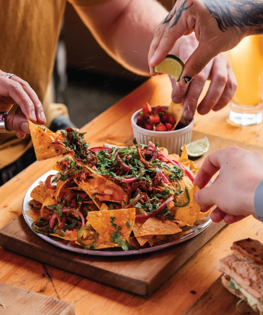 Hands grabbing meat-topped nachos with lime, cilantro, and jalapeños.