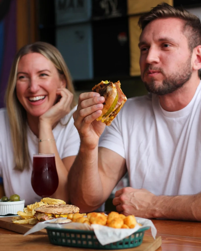 Man eats sandwich and fries as woman smiles at restaurant table.