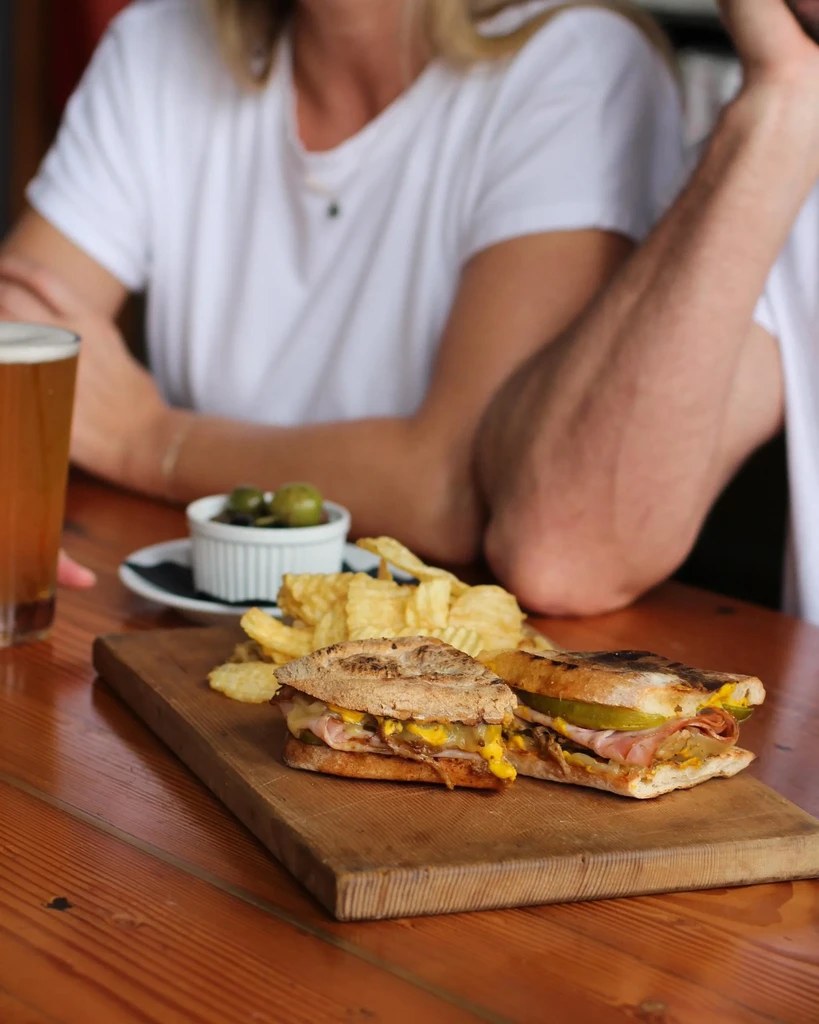 Grilled sandwich with chips, beer, and olives on a rustic wooden table.