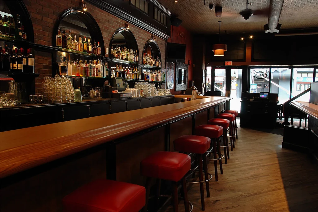 Empty bar: wooden counter, red brick wall, liquor bottles, red stools.