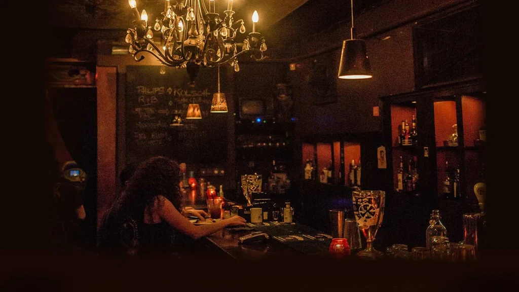 Dimly lit bar with person at counter, ornate chandelier, backlit liquor bottles.