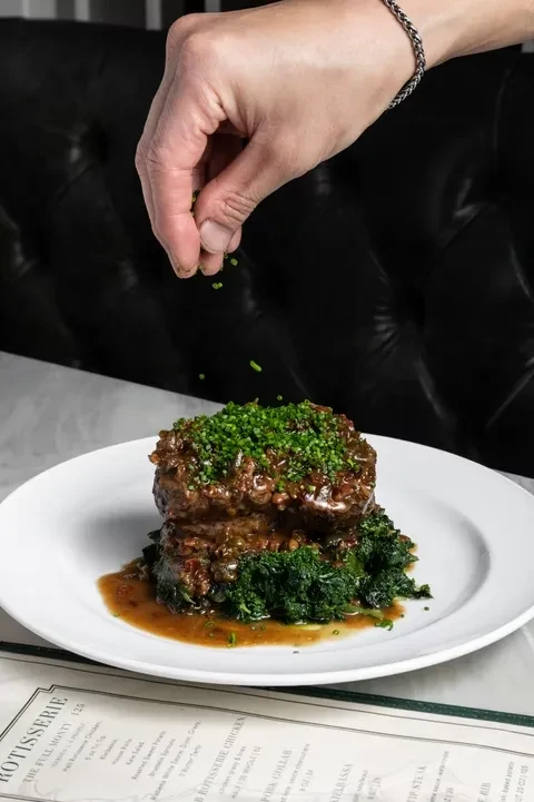 Hand garnishing plated braised meat, herbs, greens, and brown sauce.