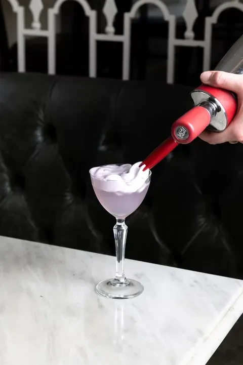 Hand pours white foam into a lavender cocktail in a stemmed glass.