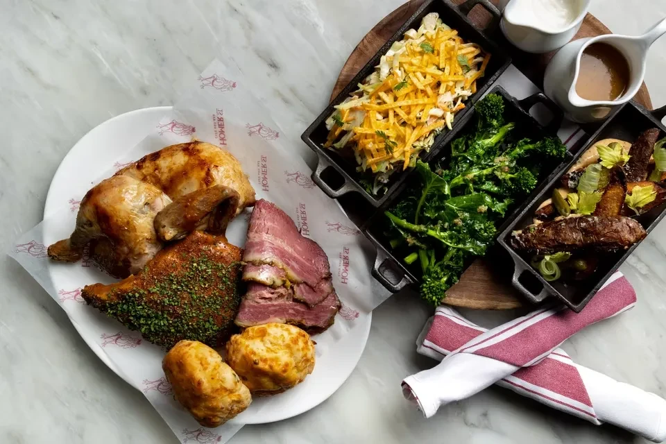 Overhead meal spread with roasted chicken, beef, vegetables, biscuits, and sauces.