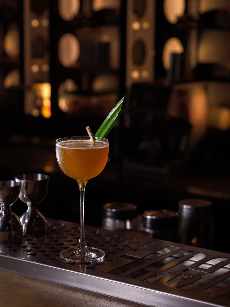 Amber cocktail in coupe glass with green leaf on metallic bar.