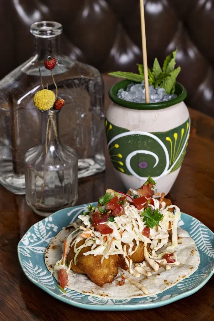 Two fish tacos, a mint cocktail, and bottles on a wooden table.