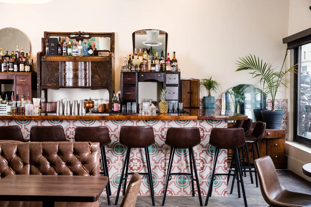 Vibrant bar with pink-teal tiled counter, antique wooden shelves, and pineapple.