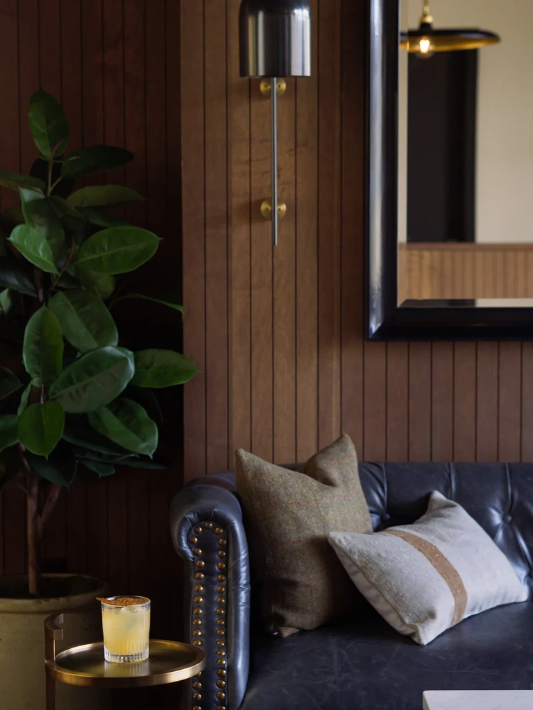 Cozy lounge with dark Chesterfield sofa, cocktail, plant, wood panels.