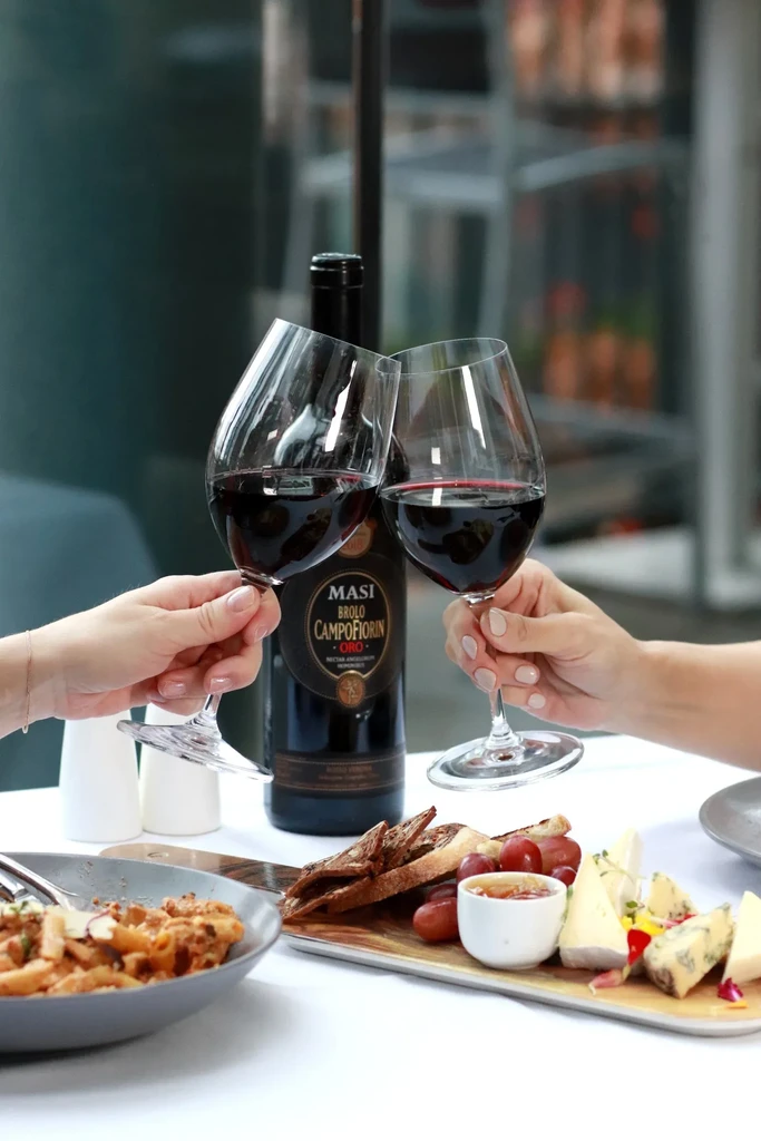 Hands toast red wine beside pasta, cheese, and a wine bottle.