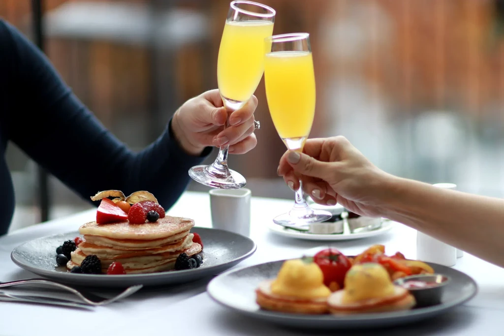 Hands toasting mimosas over pancakes and eggs benedict.