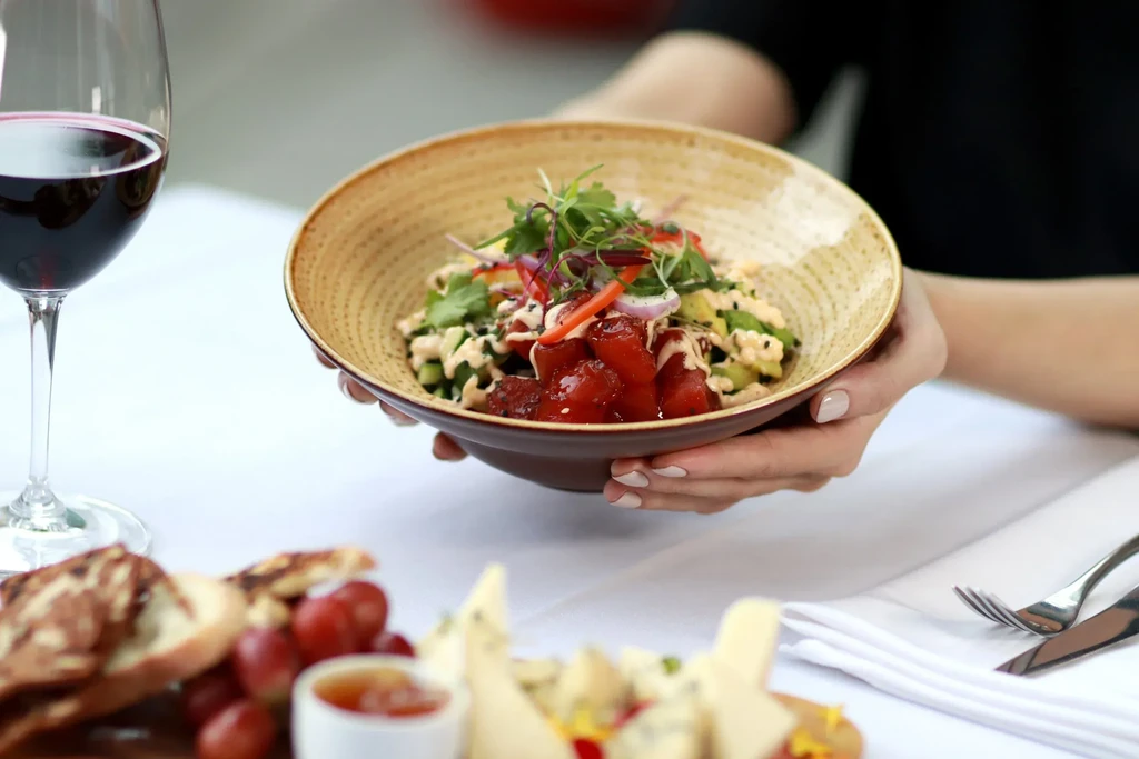 Hands holding a colorful poke bowl with wine and charcuterie.