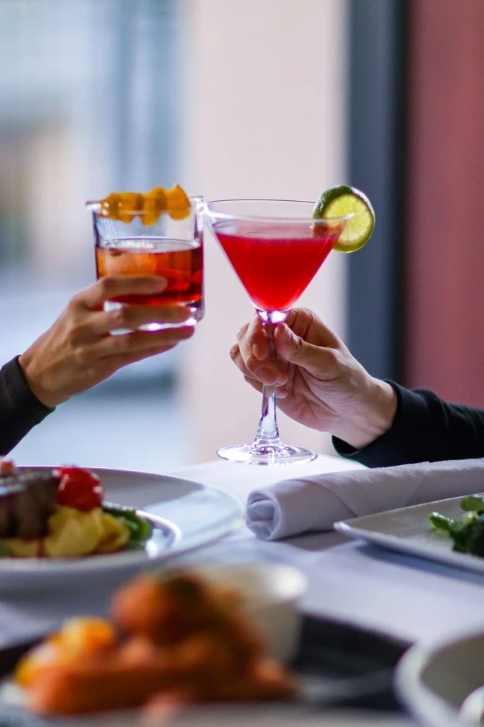 Two hands clink an amber old-fashioned and a red martini cocktail.
