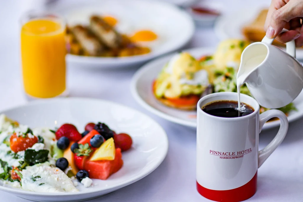 Hand pouring creamer into a Pinnacle Hotel coffee mug with breakfast.