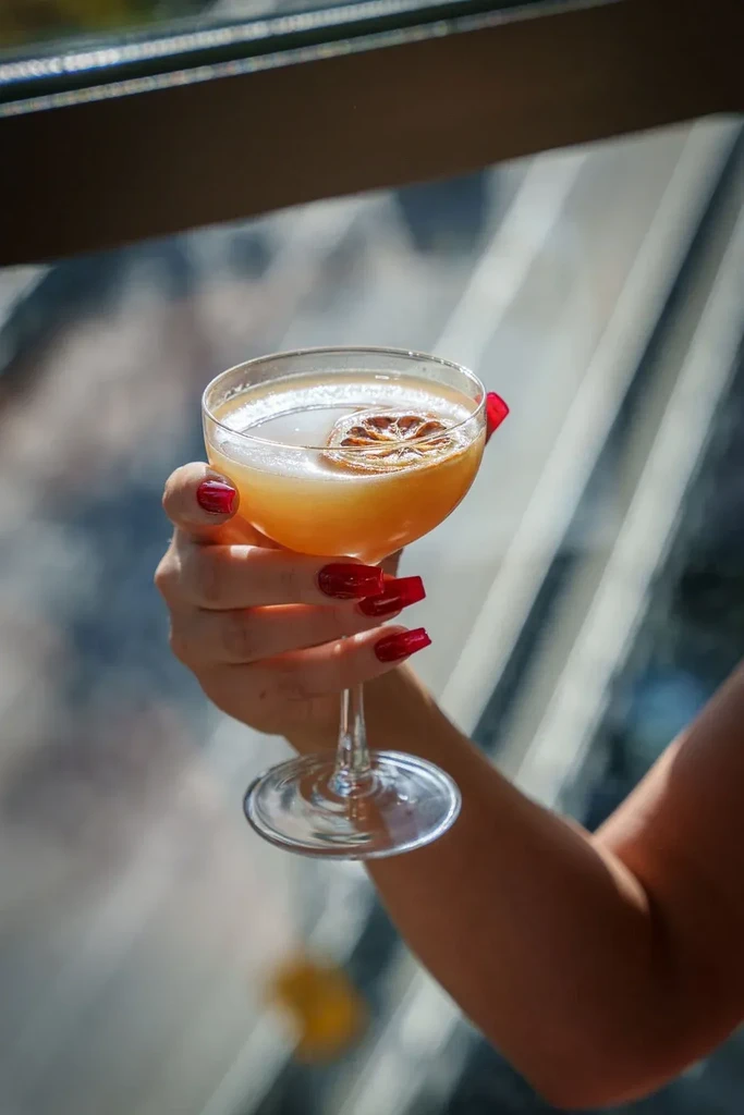 Hand with red nails holds an amber cocktail garnished with citrus.