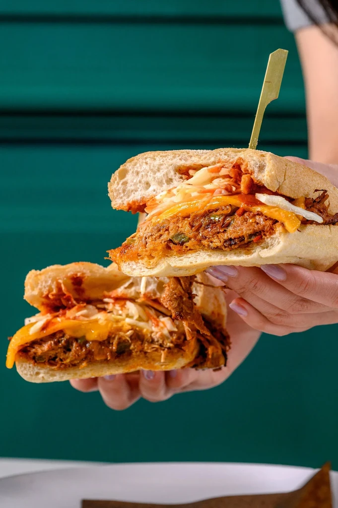 Hands hold a rustic sandwich filled with pulled meat, cheese, and slaw.