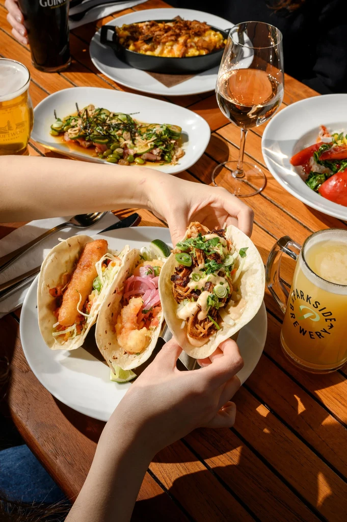 Hands holding tacos, varied food, and drinks on a sunlit wooden table.