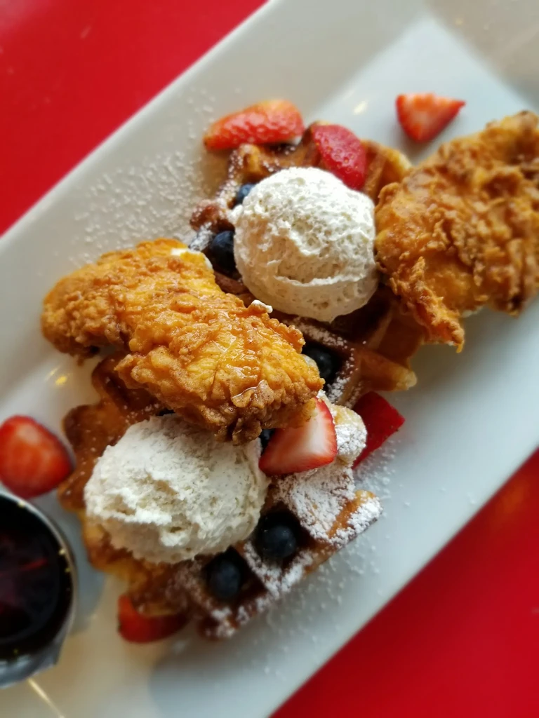 Top-down chicken and waffles with fruit, ice cream, syrup, red background.