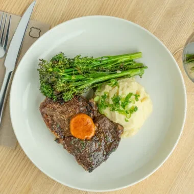 Cooked steak, mashed potatoes, broccolini, and orange sauce on white plate.