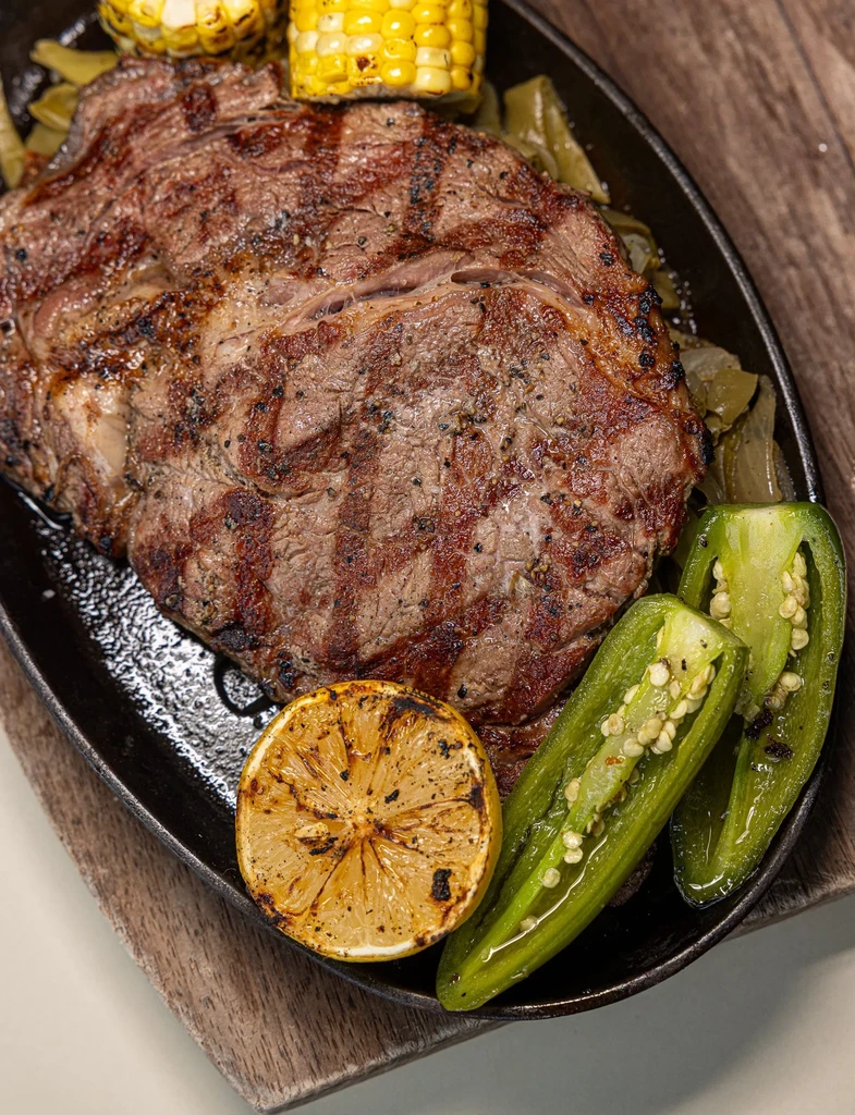 Grilled ribeye steak in skillet with grilled corn, lemon, jalapeños.