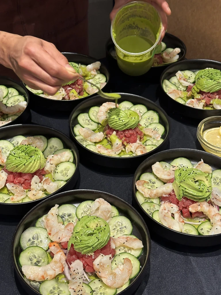 Hands preparing multiple fresh poke bowls with seafood, avocado, and sauce.