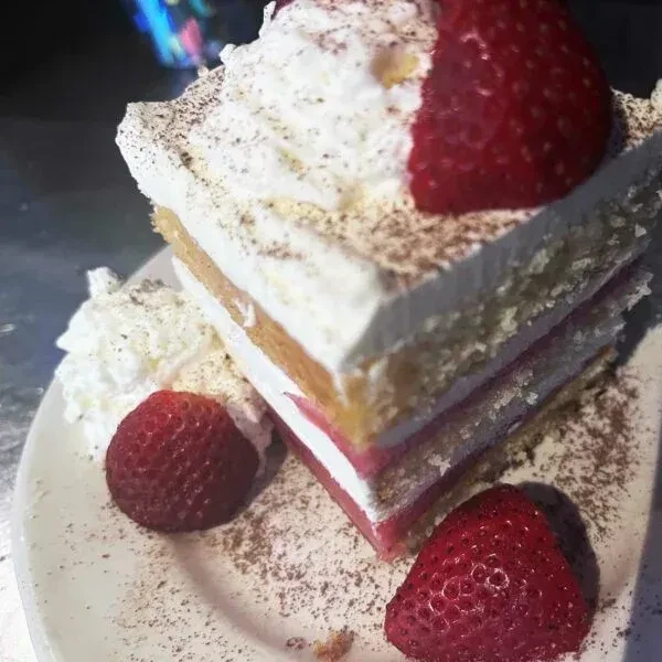 Layered cake slice with pink filling, whipped cream, strawberries, and cocoa dust.