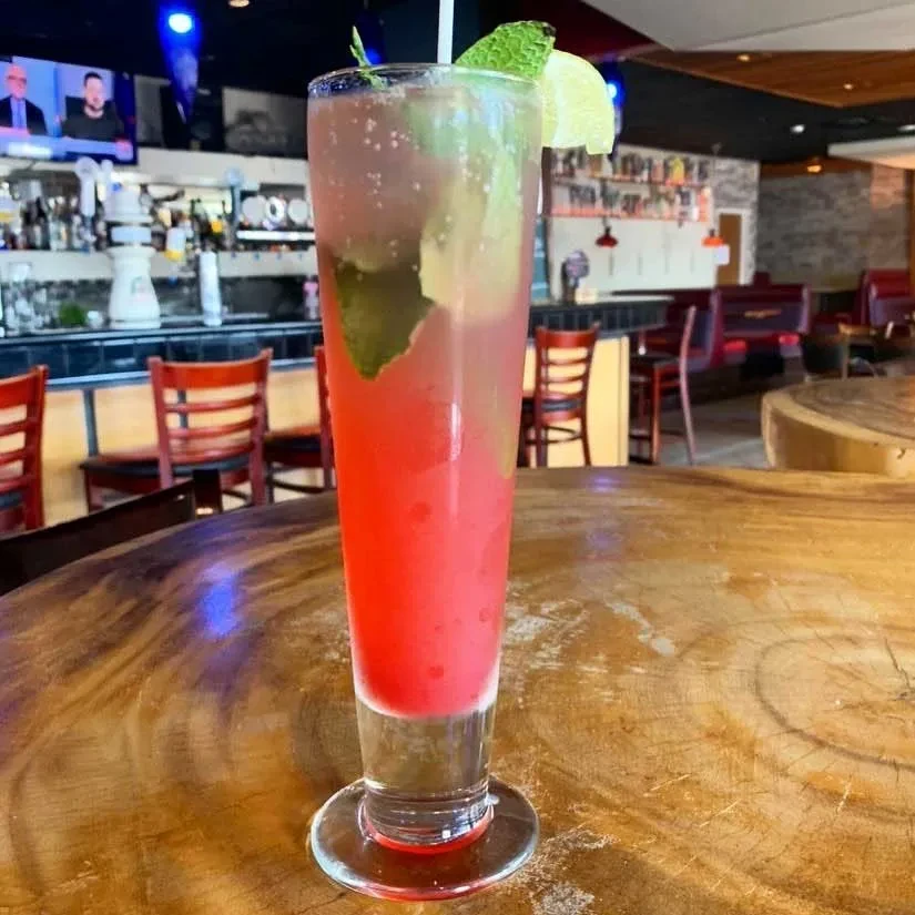 Reddish-pink drink with mint, lime, ice on table in bar.