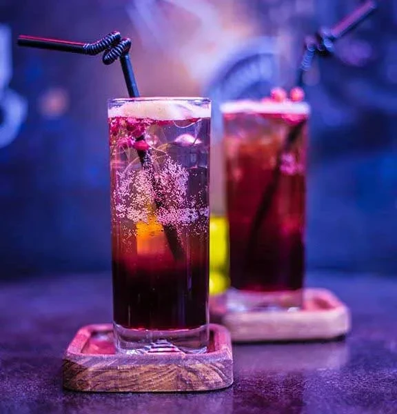 Two tall, garnished, layered drinks on dark coasters in dim light.