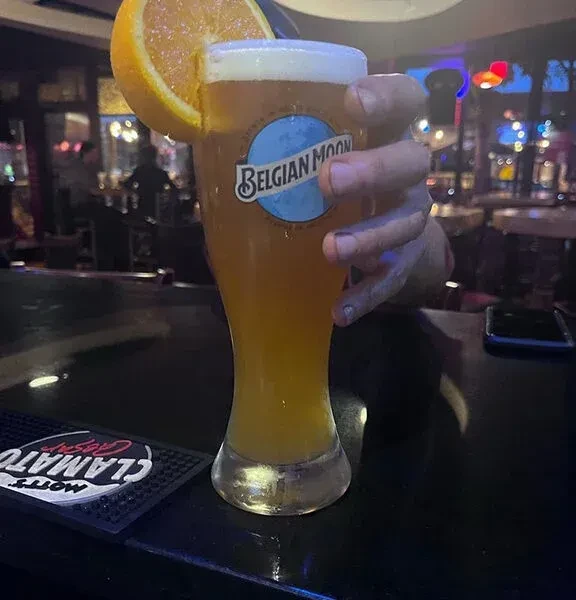 Close-up amber Belgian Moon beer with orange slice, held on dark bar.