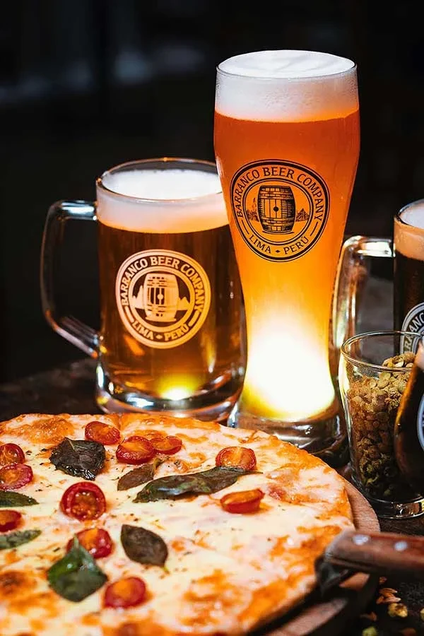 Close-up of pizza with cherry tomatoes and basil, next to Barranco beer glasses.