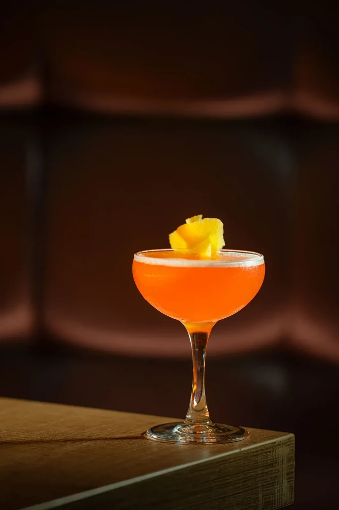 Vibrant orange cocktail with citrus peel in coupe on wood.