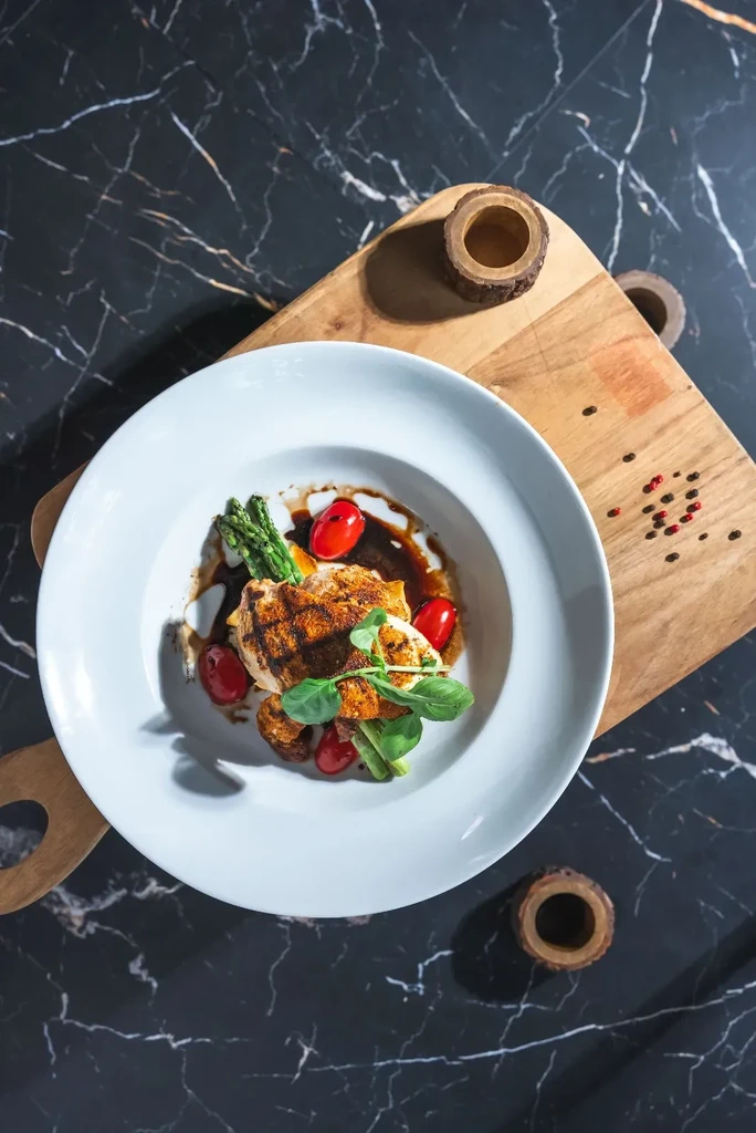 Plated grilled protein, asparagus, tomatoes, and sauce on a wooden board.